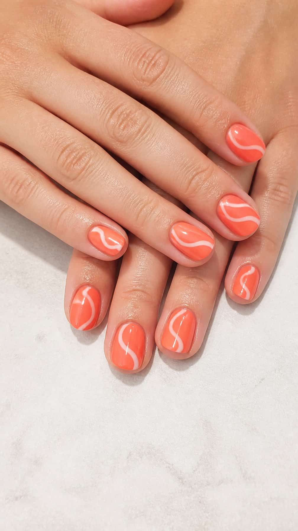 A close-up of coral-colored nails decorated with simple white wave designs.