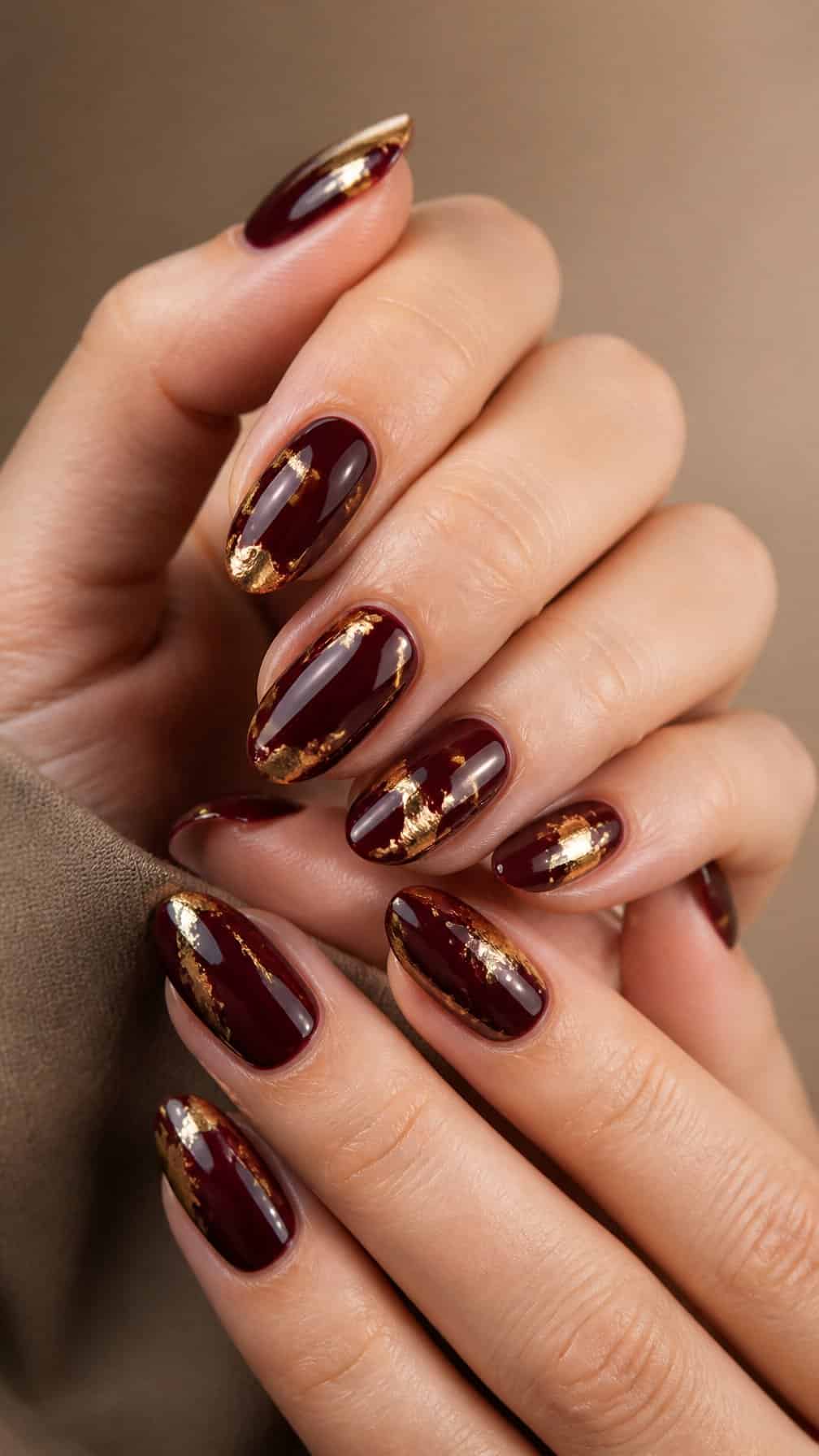 A close-up of manicured hands with glossy deep burgundy nails accented with gold foil.