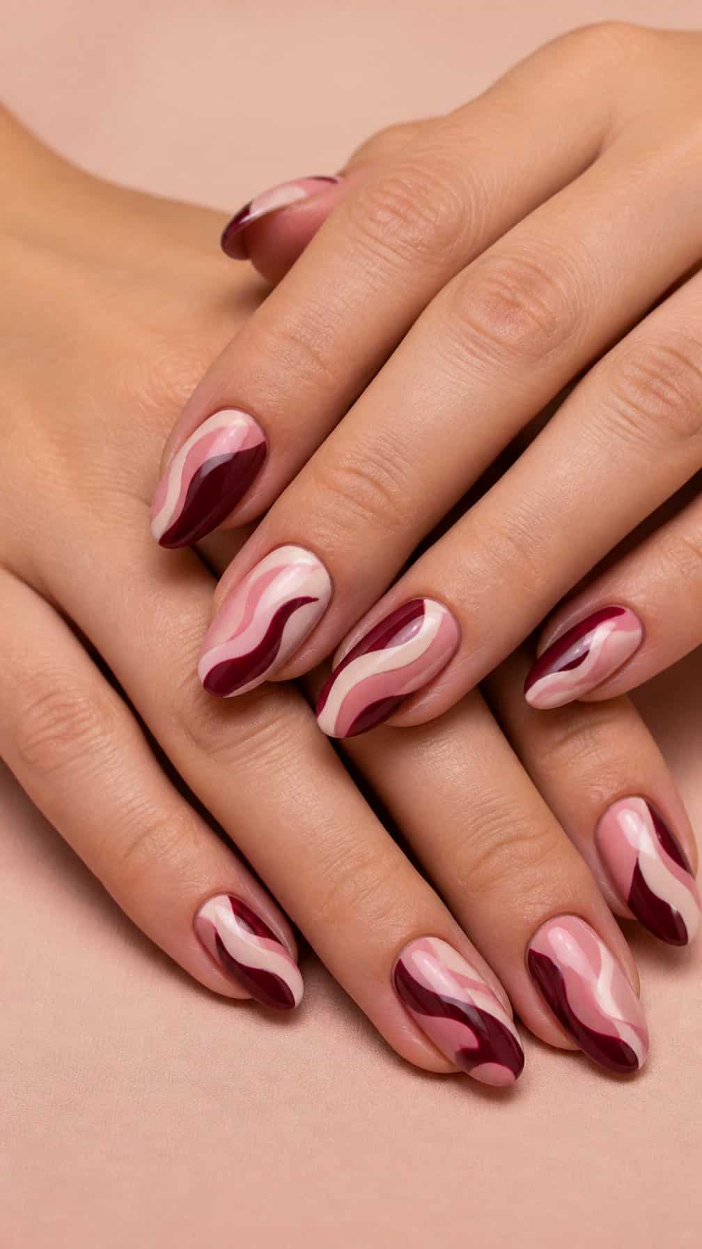 A close-up of hands with maroon and blush pink swirl nail designs.