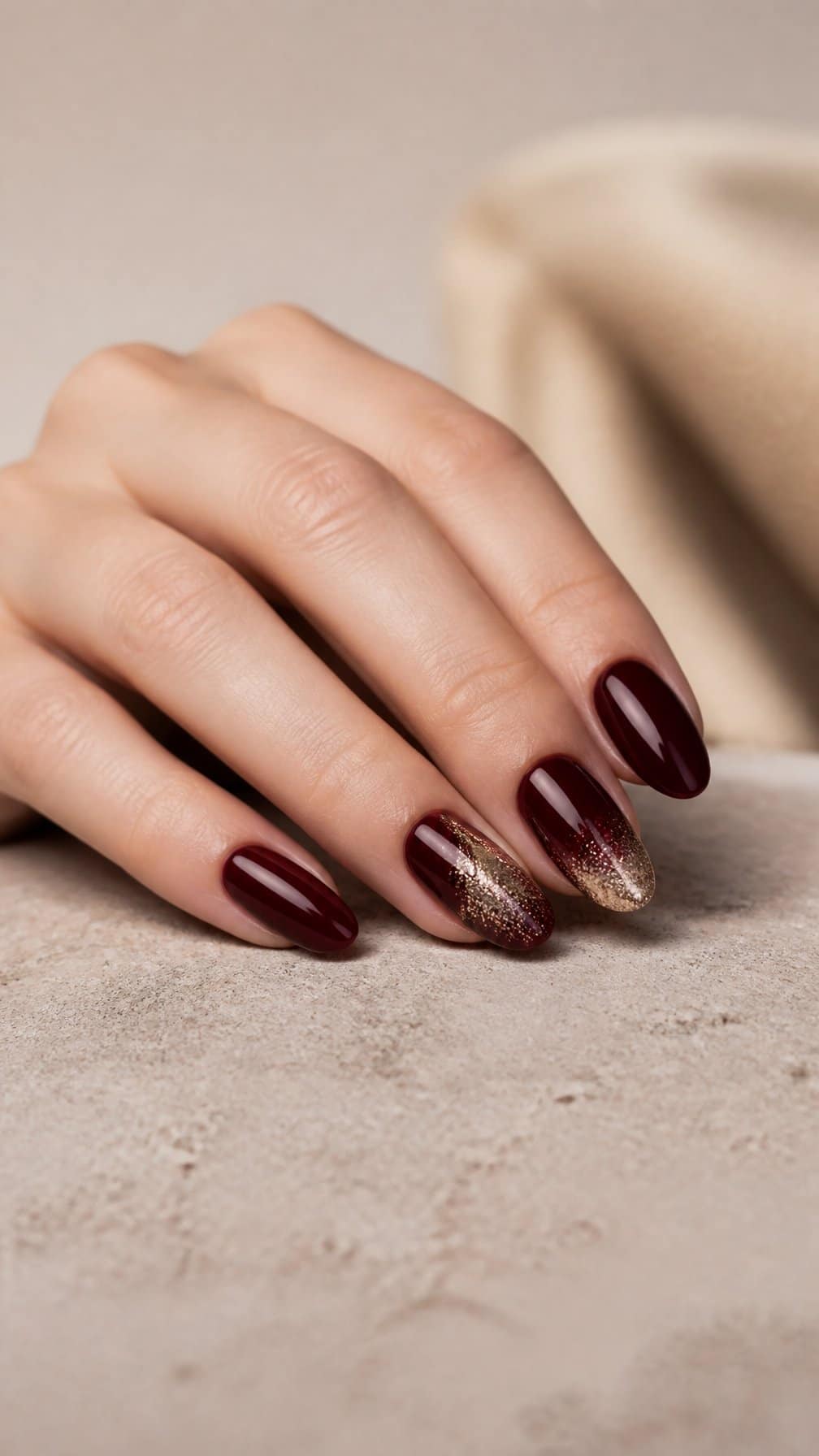 A manicured hand with glossy maroon nails and champagne-gold glitter accents resting on a neutral surface.