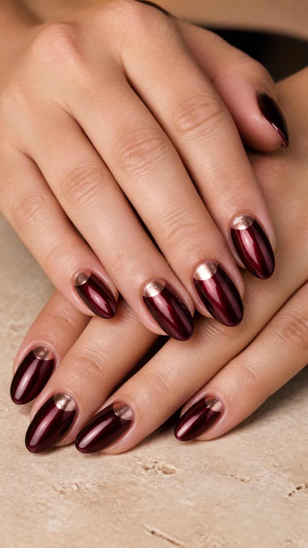 A hand displays glossy maroon nails with metallic half-moon accents.