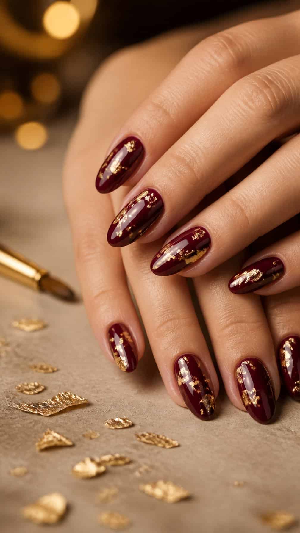 A hand with glossy maroon almond-shaped nails decorated with gold foil flakes rests on a neutral surface.
