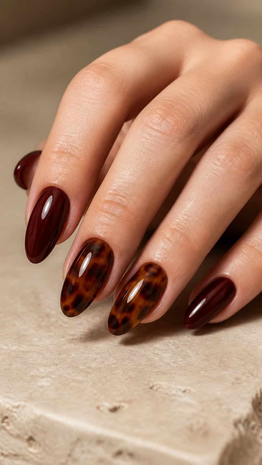 A hand with glossy maroon nails and tortoiseshell accent nails rests on a beige surface.
