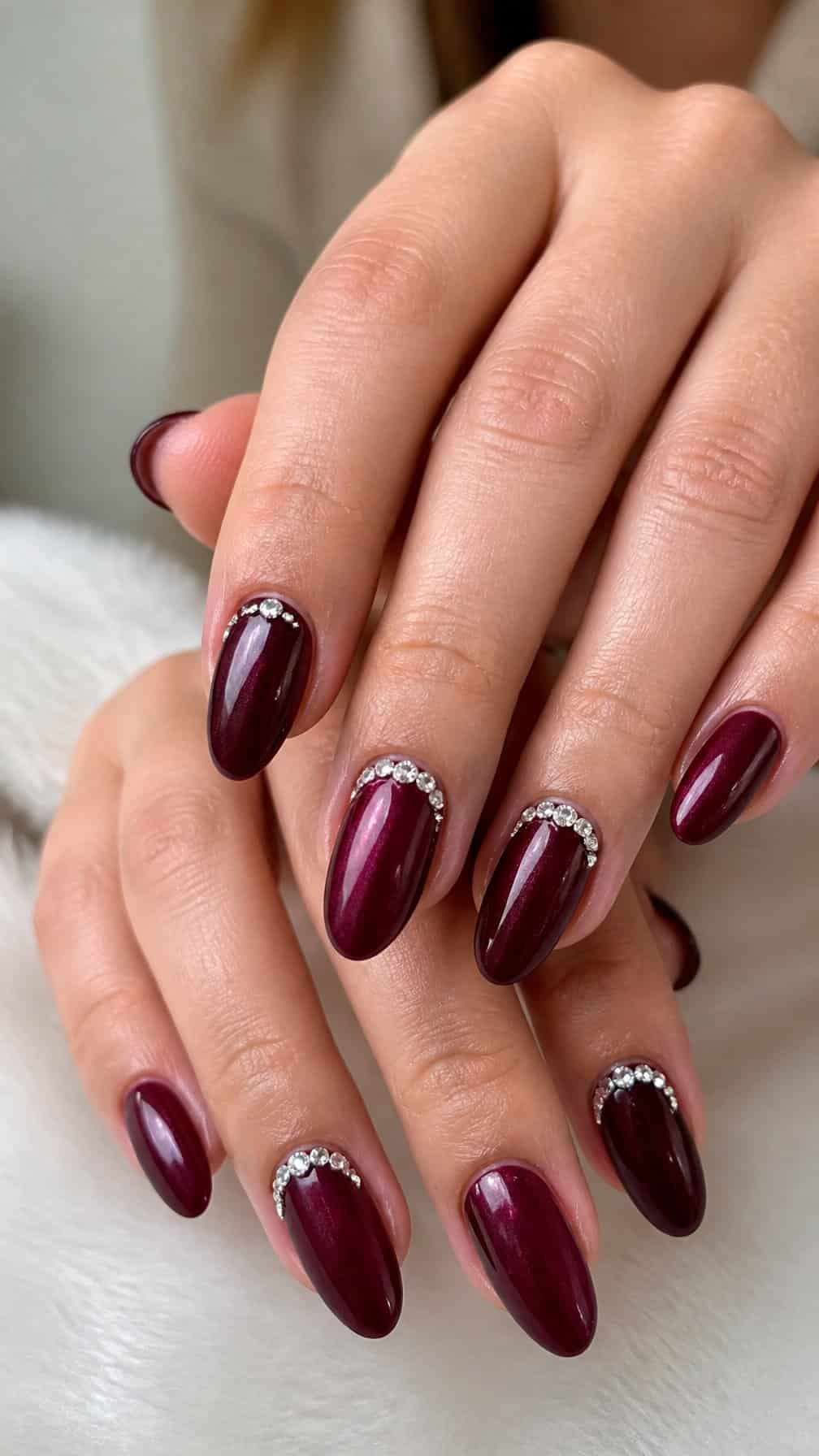 Close-up of hands with deep merlot-colored almond-shaped nails decorated with small rhinestones.