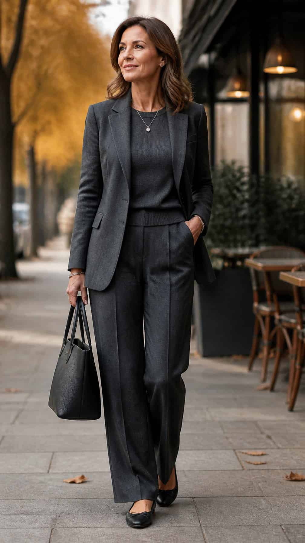 A confident woman wearing a monochromatic charcoal outfit with ballet flats stands outdoors among autumn foliage.