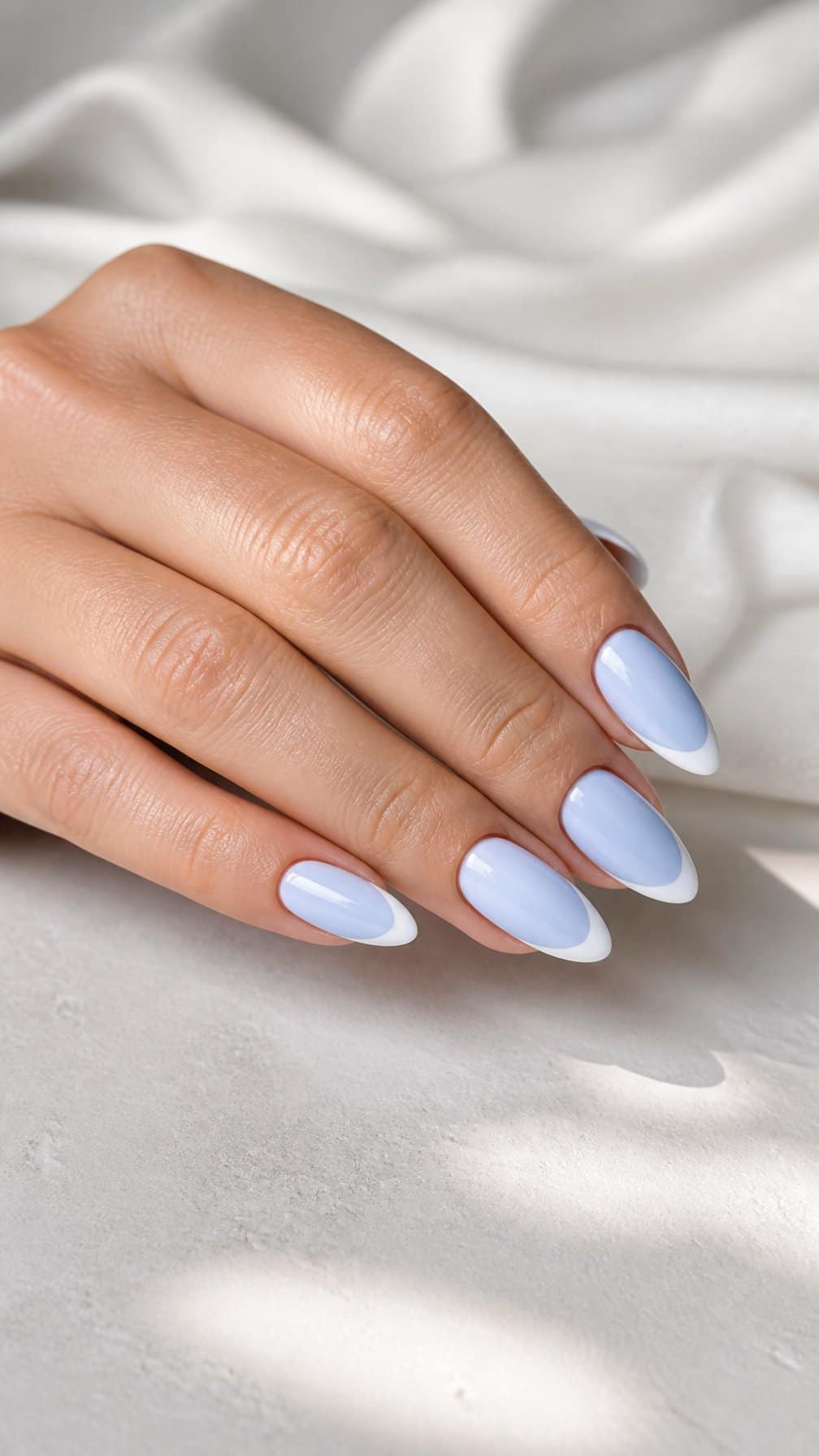 A hand with glossy pastel blue nails featuring white French tips rests on a light neutral surface.
