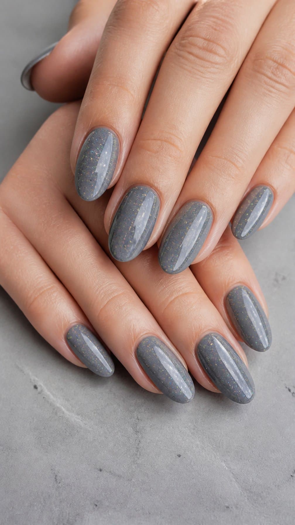 A close-up of hands with slate gray holographic nails resting on a light gray surface.