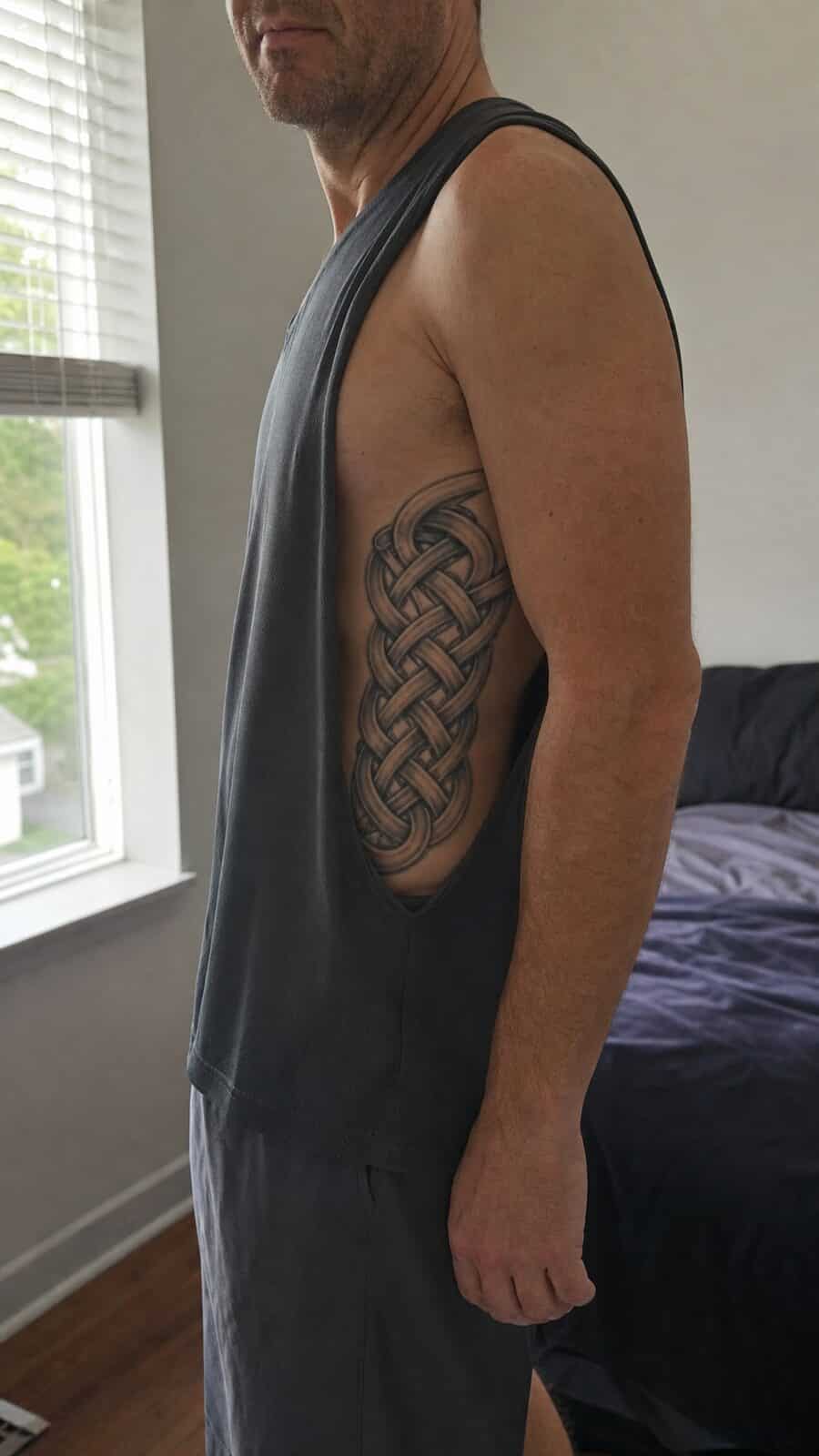 25 Rib Tattoos for Men Worth Considering 17 Healed celtic knot tattoo on a adult man's side ribs, photographed in a natural waist-height candid close-up — study 1