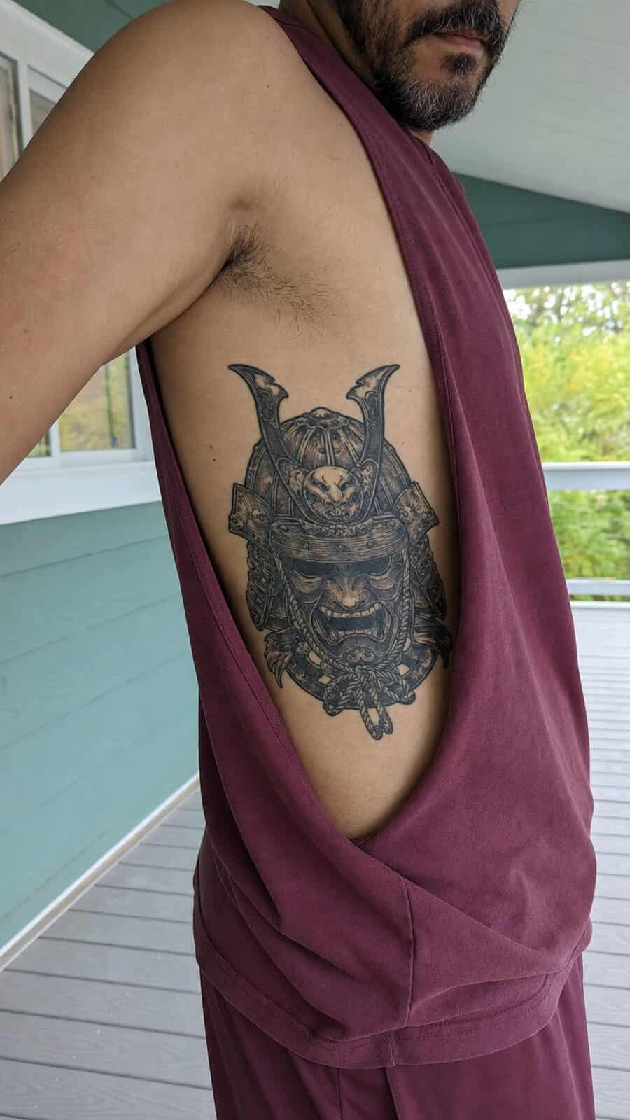 25 Rib Tattoos for Men Worth Considering 30 Healed samurai mask tattoo on a adult man's side ribs, photographed in a natural slightly elevated close view — study 1