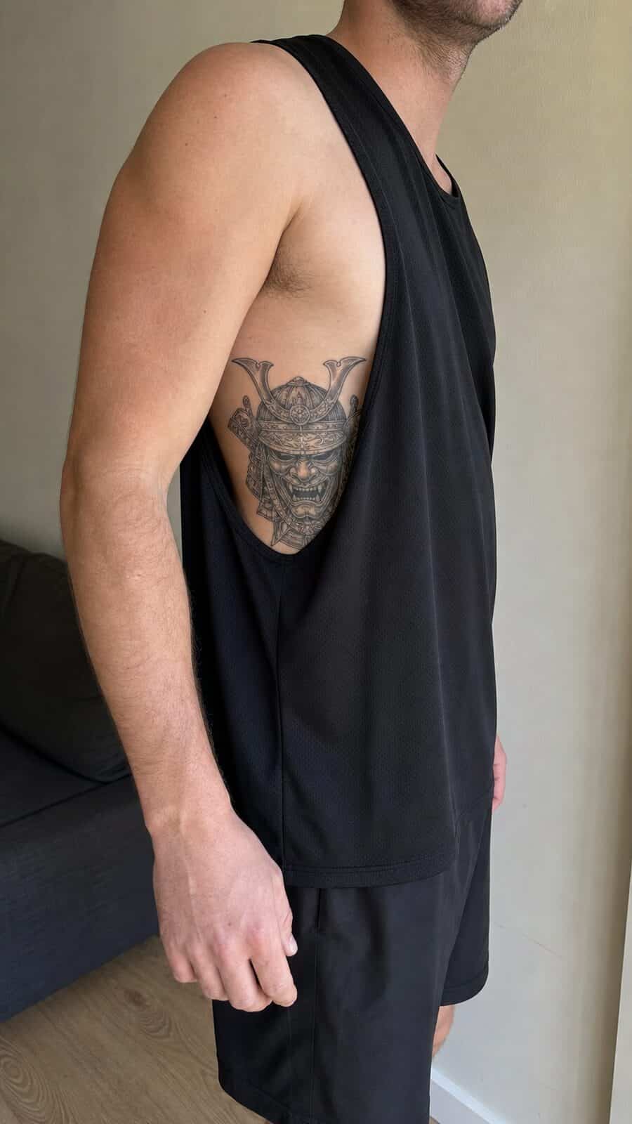 25 Rib Tattoos for Men Worth Considering 31 Healed samurai mask tattoo on a adult man's side ribs, photographed in a natural waist-height candid close-up — study 2