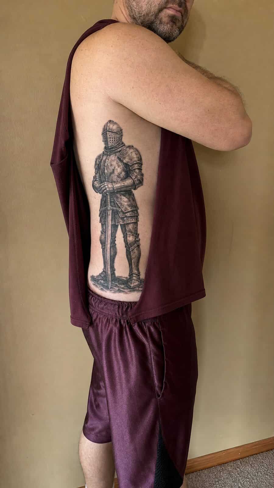 25 Rib Tattoos for Men Worth Considering 51 Healed knight in armor tattoo on a adult man's side ribs, photographed in a natural slightly elevated close view — study 1