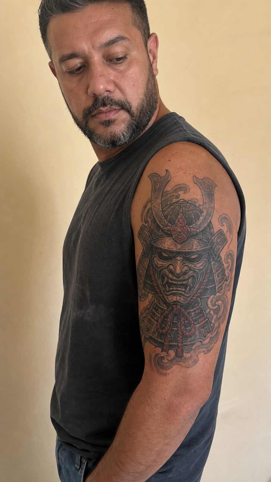 Unlock Your Next Ink: 25 Epic Upper Arm Tattoo Ideas for Men 62 Healed samurai mask tattoo on a adult man's outer upper arm, photographed in a natural relaxed documentary crop — study 2