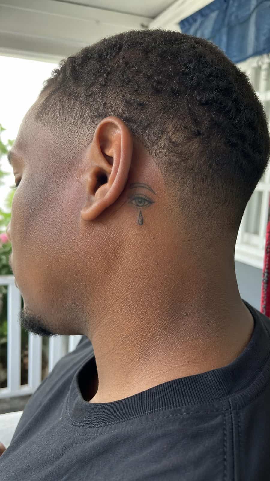 Healed tearful eye tattoo tattoo on a adult person's behind the ear, photographed in a natural casual close three-quarter view — study 1