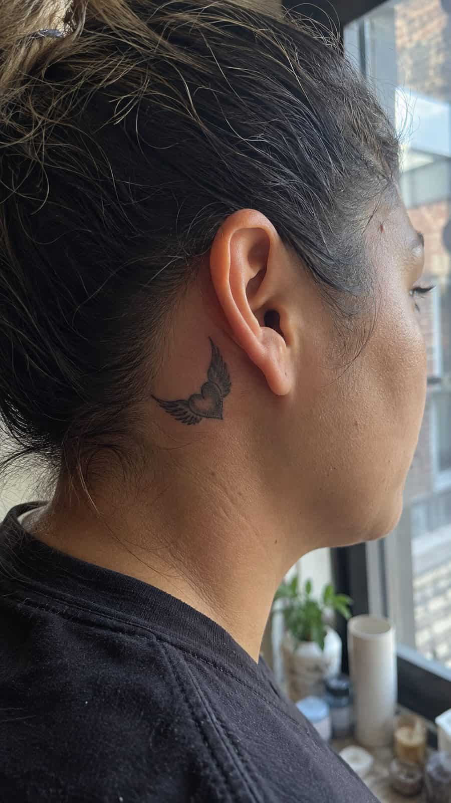 Healed winged heart tattoo on a adult person's behind the ear, photographed in a natural casual close three-quarter view — study 1