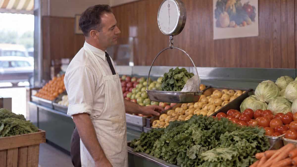 15 Grocery Store Features from the '60s That Have Vanished 15 Grocery clerk weighing produce on a hanging scale.