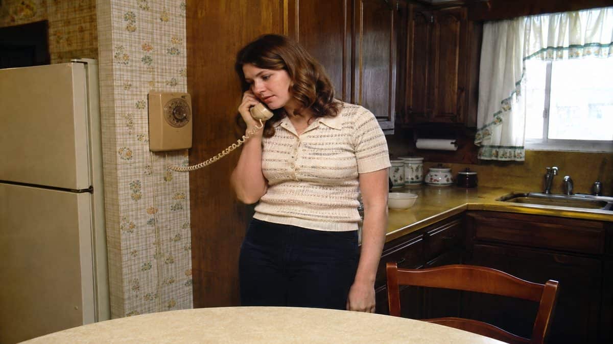 Woman talking on a rotary wall phone in a kitchen.