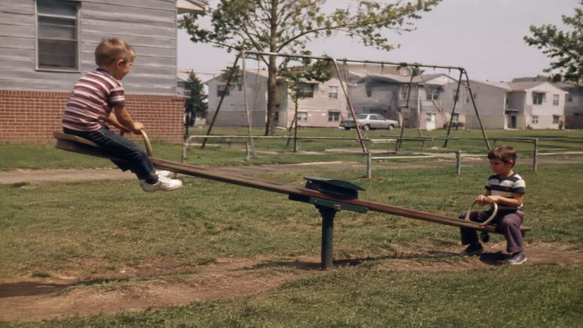 Remember These? 15 Playground Equipment Pieces That Would Never Pass Safety Standards Today 6 Real vintage photo of kids playing on a wooden or metal see-saw.