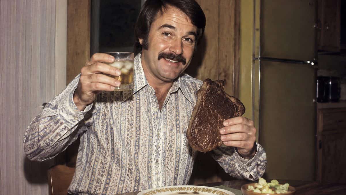 Vintage 1970s photo of a man cheerfully holding a steak and a drink.