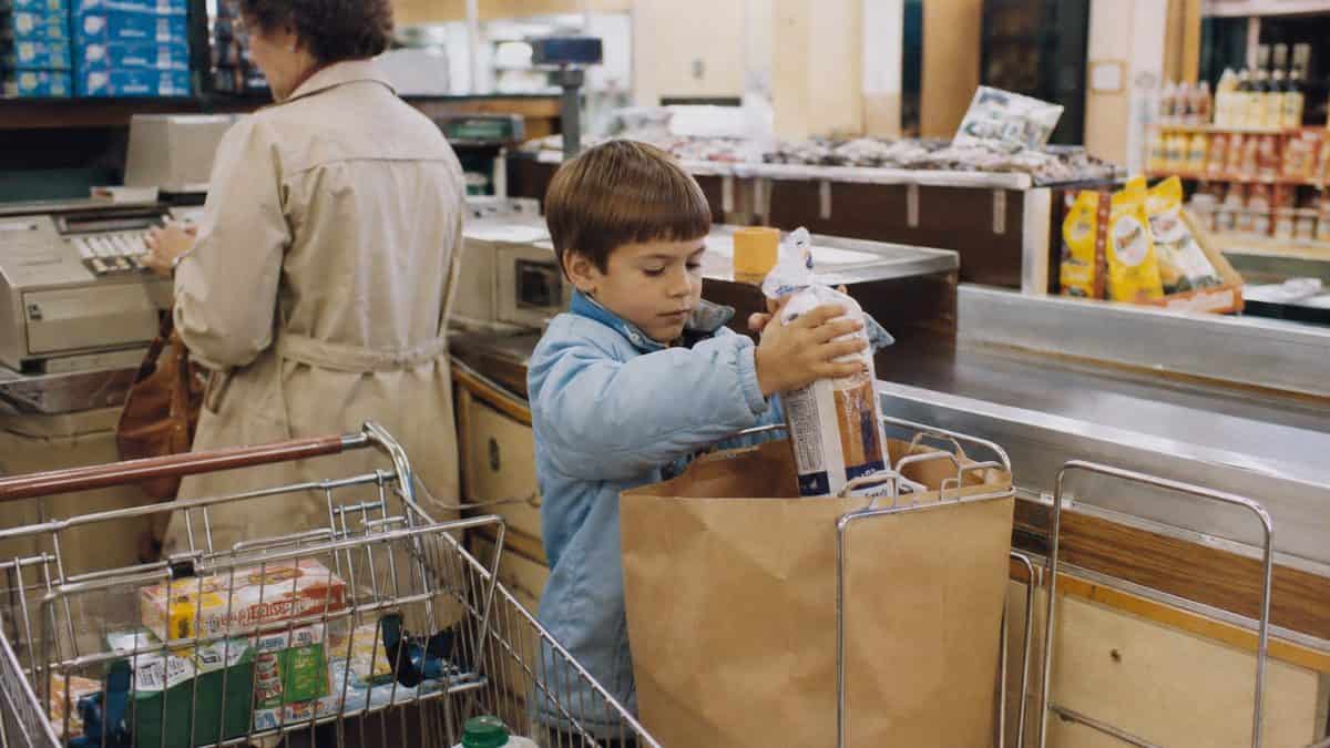 What Kids Used to Do at the Grocery Store That's Unthinkable Now 5 Child helping bag groceries at checkout with natural human features and realistic, readable brand packaging