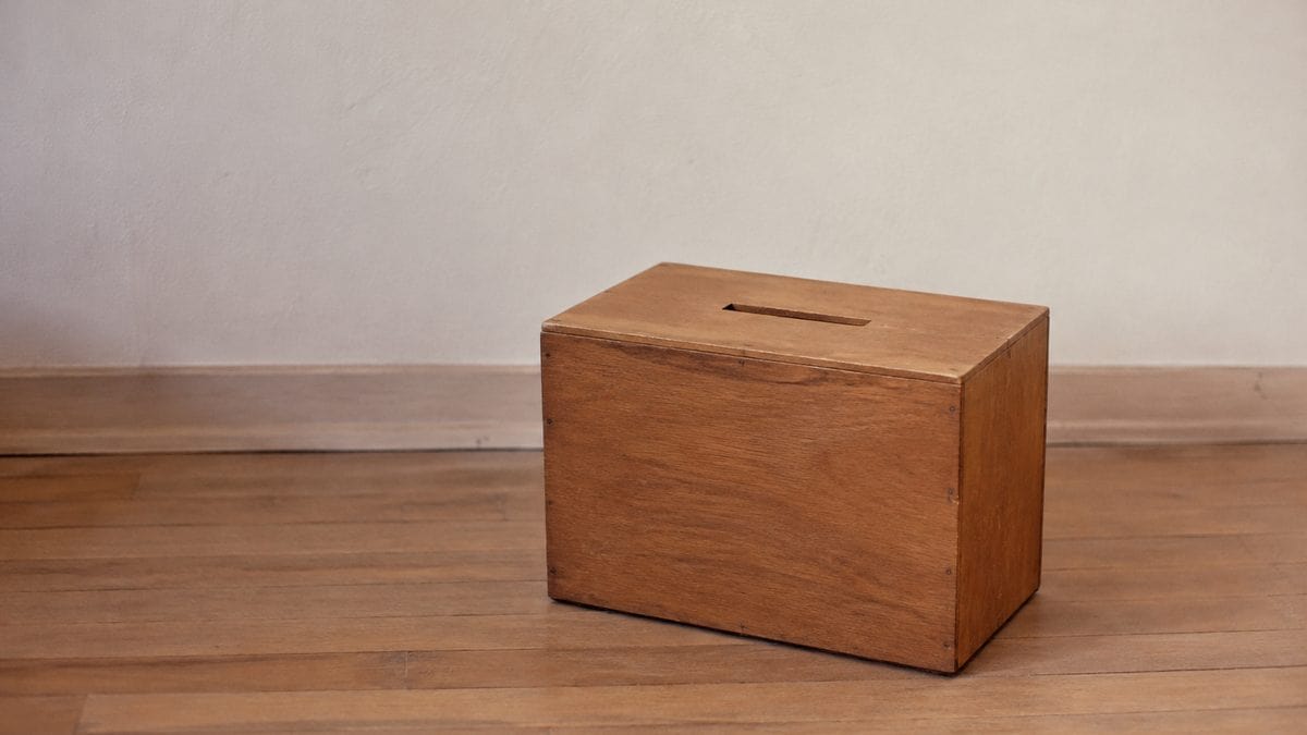 Wooden classroom ballot box with a clearly cut rectangular slot on top, alone against a plain wall
