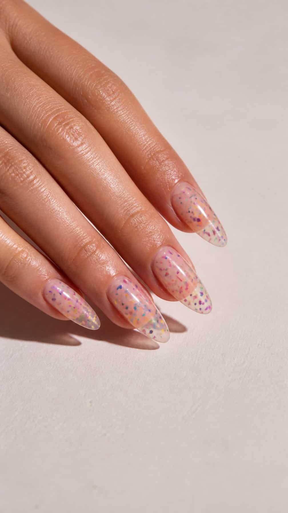 A hand with long clear almond-shaped nails decorated with colorful confetti glitter.