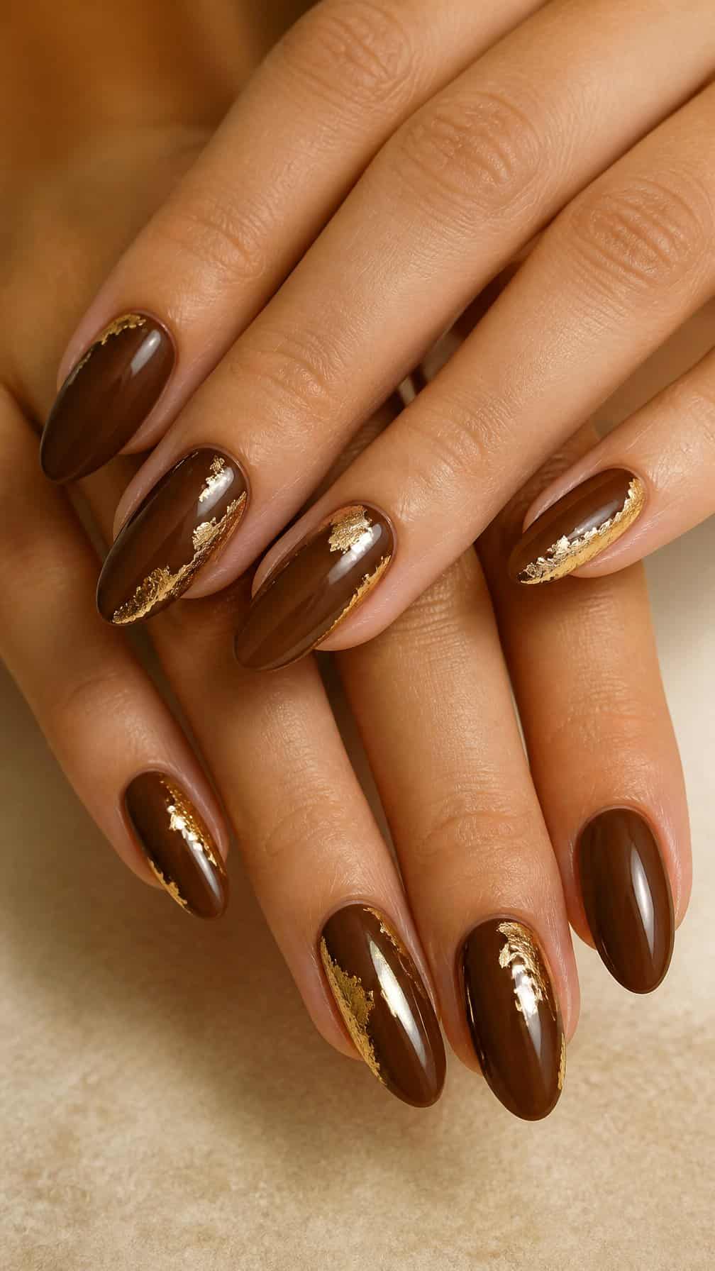 A close-up of almond-shaped walnut brown nails with delicate gold leaf accents.