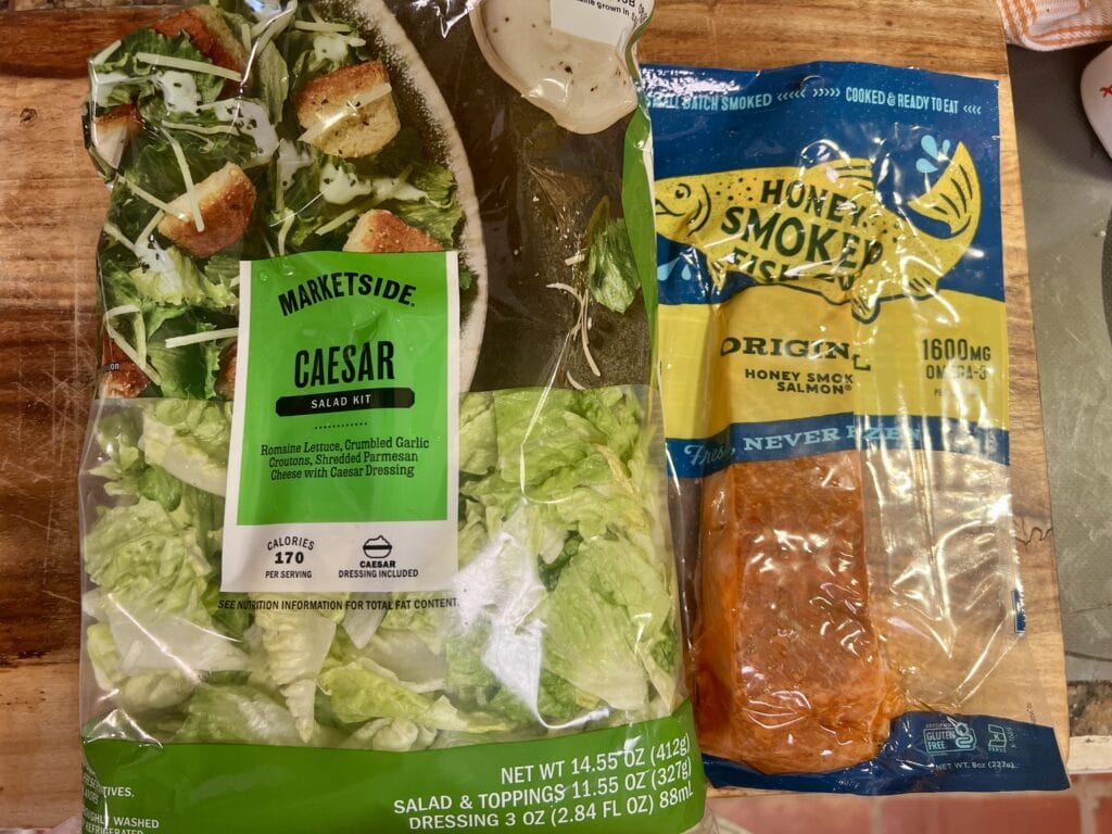 Fresh Caesar salad kit and smoked salmon package on wooden surface.