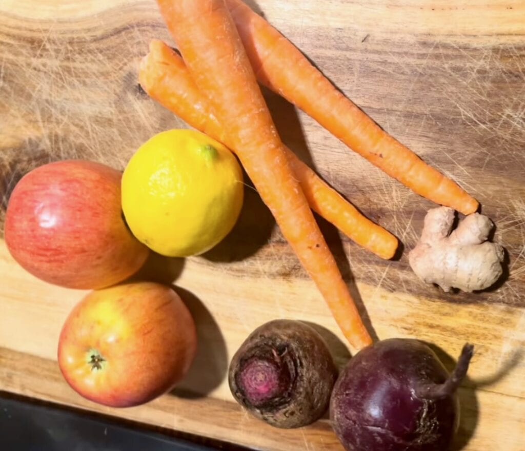 Fresh carrots, apples, lemon, ginger, and beets on a wooden cutting board.