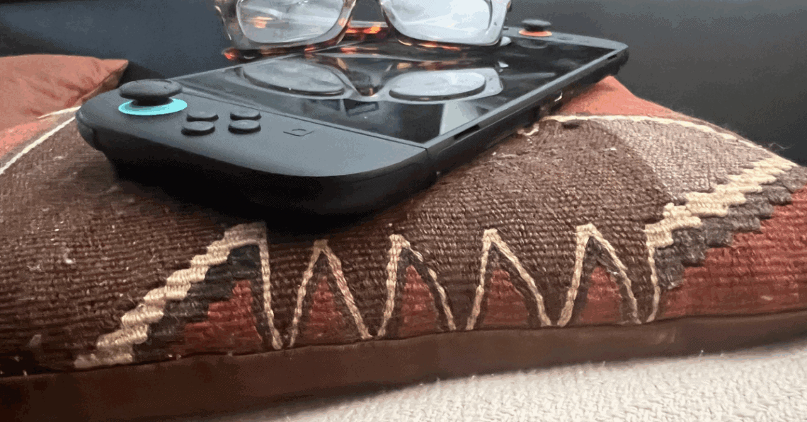 Close-up of a handheld gaming console with glasses resting on top, placed on a patterned pillow with earth tones and stitched detailing.