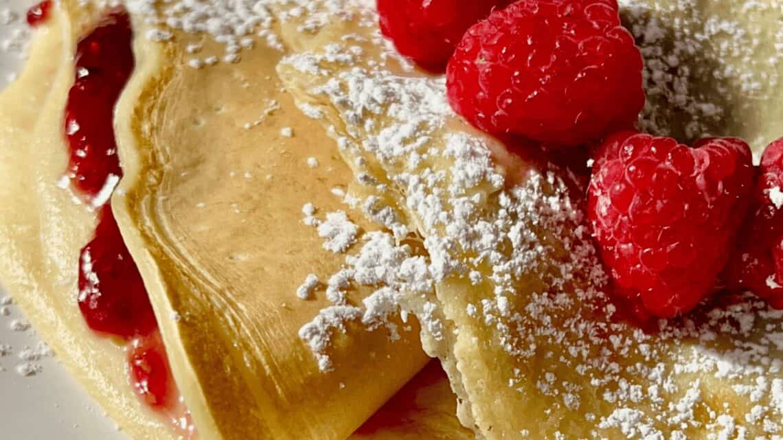 Freshly made crepes filled with raspberry jam, dusted with powdered sugar, and topped with fresh ras.