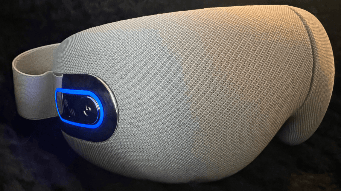 Close-up side view of a beige heated eye massager with a blue illuminated power button resting on a dark surface.