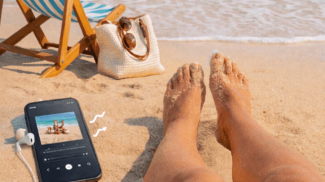 Person's feet on the sand with a smartphone, sunglasses, and a bag nearby.