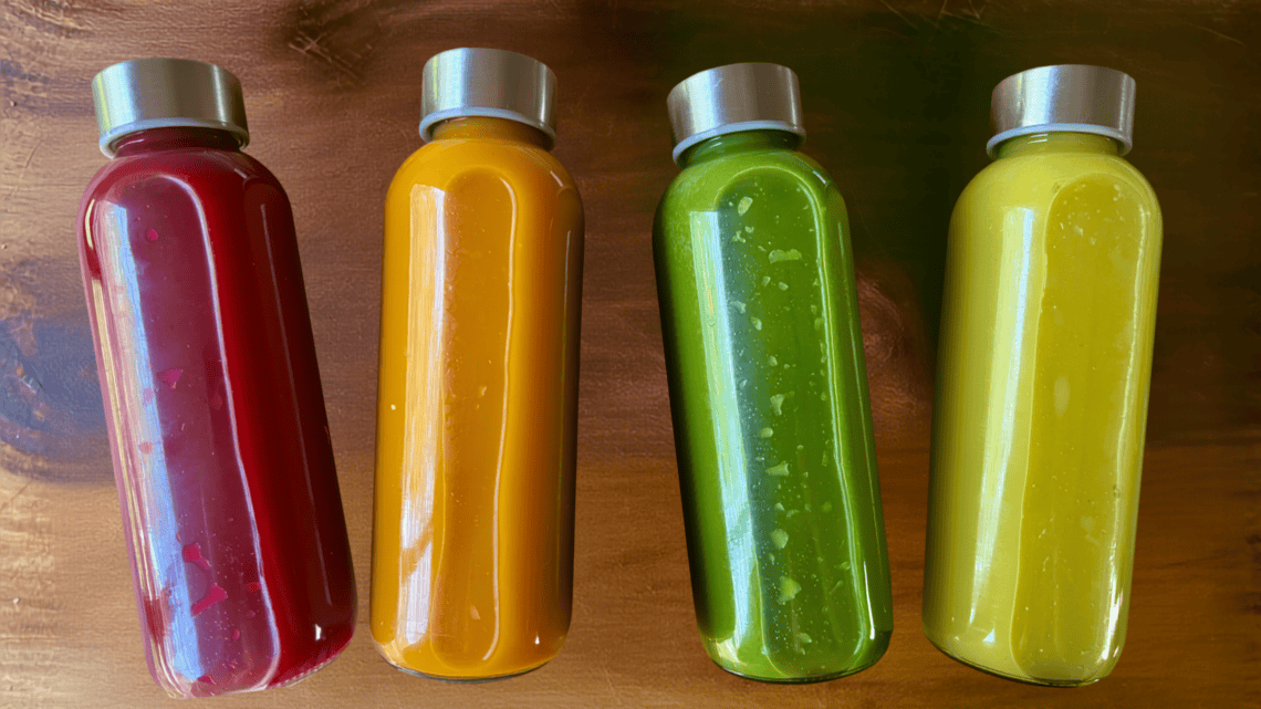 Four vibrant smoothie bottles in red, orange, green, and yellow with silver caps.