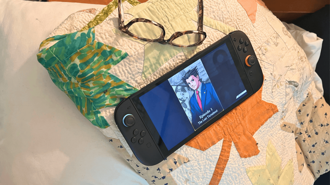 Gaming console displaying anime scene with glasses on quilted bedspread.