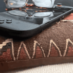 Close-up of a handheld gaming console with glasses resting on top, placed on a patterned pillow with earth tones and stitched detailing.