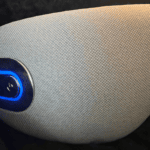 Close-up side view of a beige heated eye massager with a blue illuminated power button resting on a dark surface.