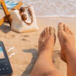 Person's feet on the sand with a smartphone, sunglasses, and a bag nearby.