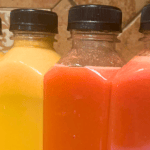 Four bottles of juice or smoothie with black caps in yellow, orange, and pink colors.