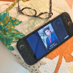 Gaming console displaying anime scene with glasses on quilted bedspread.