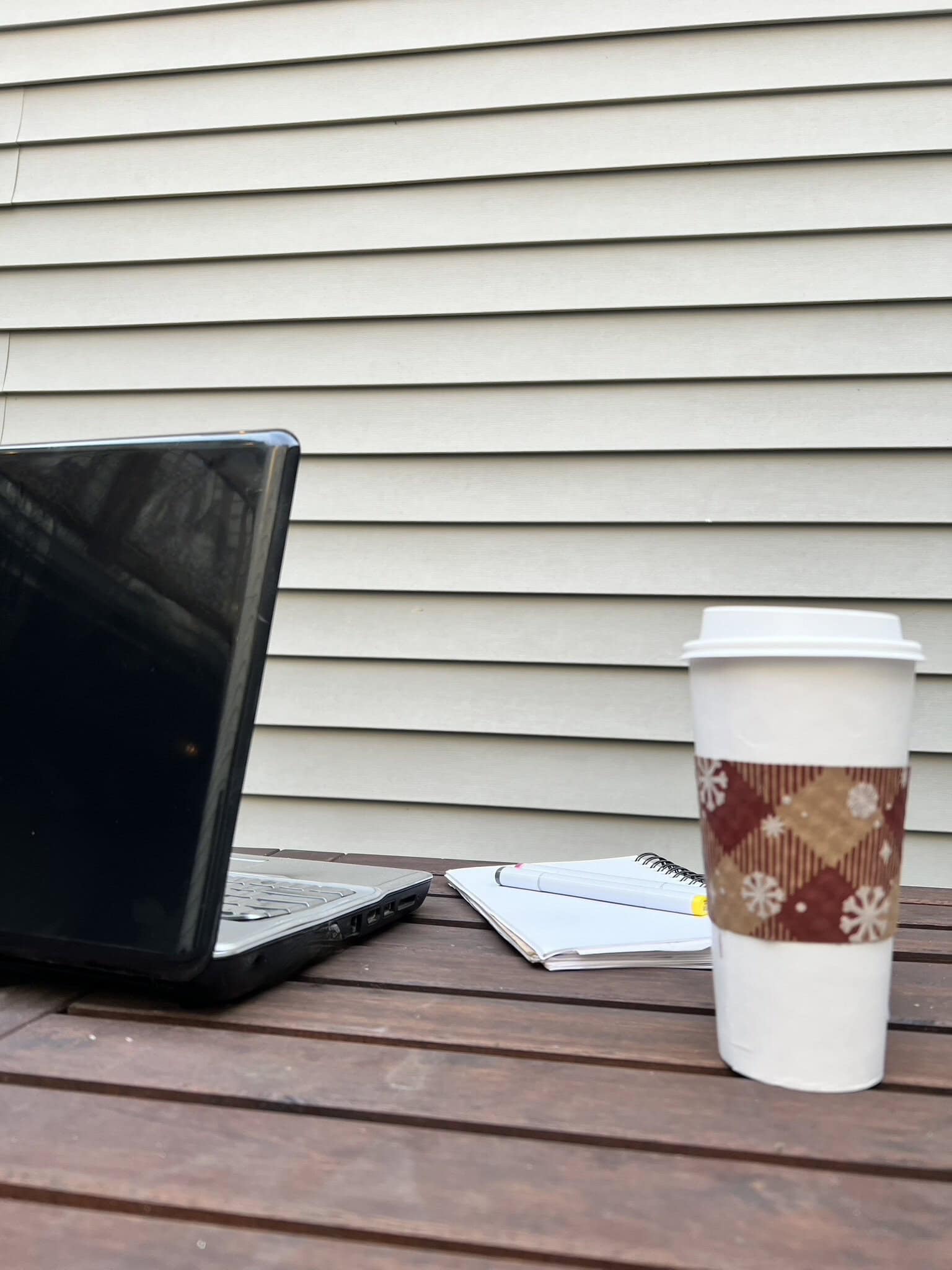 Laptop, notebook, and coffee cup on wooden table outside.