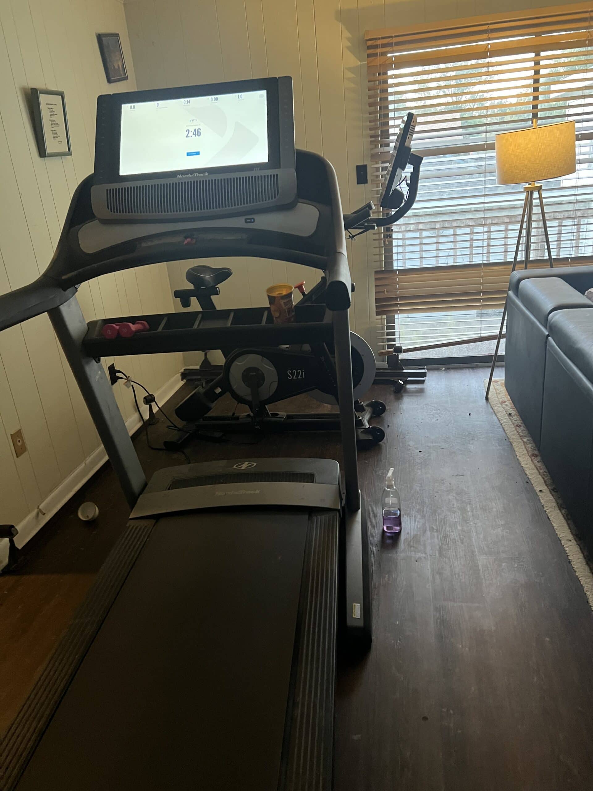 Treadmill in a cozy home gym with window blinds and a lamp.