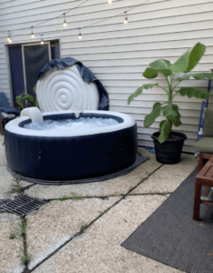 Outdoor hot tub with cover and string lights on a patio with plants and furniture.