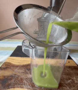 Green juice being poured through a strainer into a glass container.