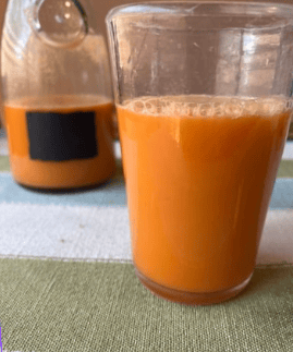 Fresh carrot juice in a clear glass with a bottle of juice in the background.