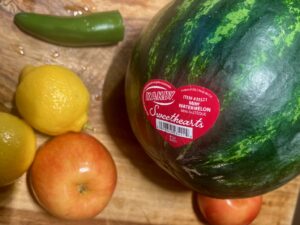 Watermelon, apples, lemons, and a jalapeño on a wooden surface.
