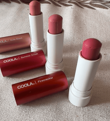 Three Coola Mineral Liplux tinted lip balms in Nude Beach, Summer Crush, and Firecracker displayed open on a neutral fabric surface with matching red and pink tubes beside them.