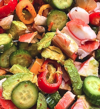 Close-up of a fresh seafood salad with imitation crab, sliced cucumbers, avocado chunks, and colorful mini sweet peppers tossed together.