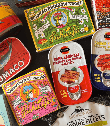 Assorted colorful tins of smoked seafood including rainbow trout, mackerel, sardines, and boneless butterflied brisling arranged flat lay style on a dark surface.