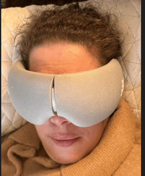 Woman lying down wearing a beige heated eye massager mask, wrapped in a tan blanket during a relaxing self-care session.