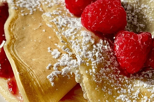 Freshly made crepes filled with raspberry jam, dusted with powdered sugar, and topped with fresh ras.
